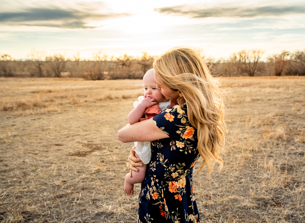 mom holding and kissing baby while her hair blows in the wind standing in an open field with a soft sunset