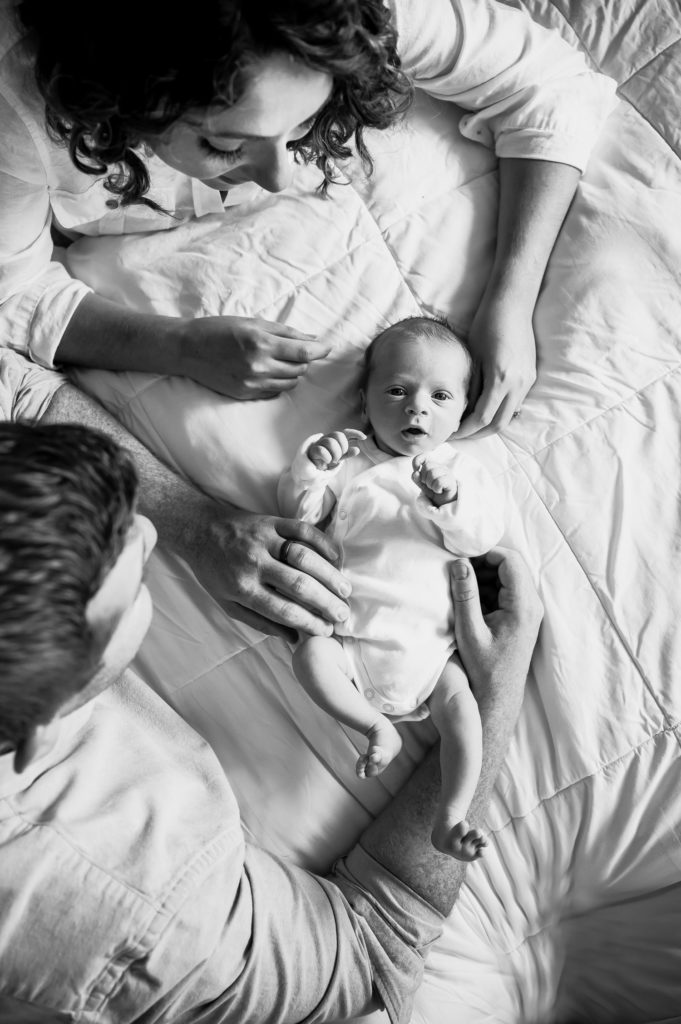 black and white picture of baby laying on bed looking at camera while parents look at him

