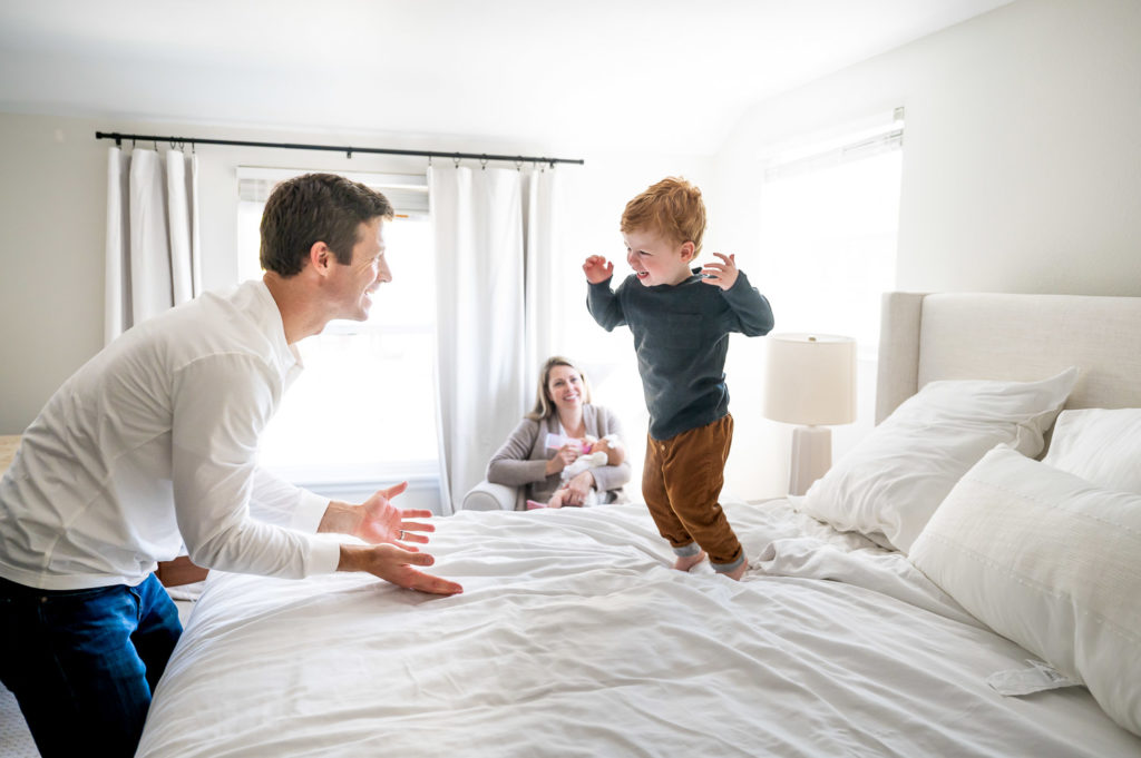 big brother jumping on bed with dad while mom and baby watch
