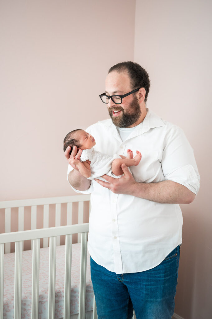 Dad holding newborn baby in front of crib in a pink nursery in Wash Park Denver