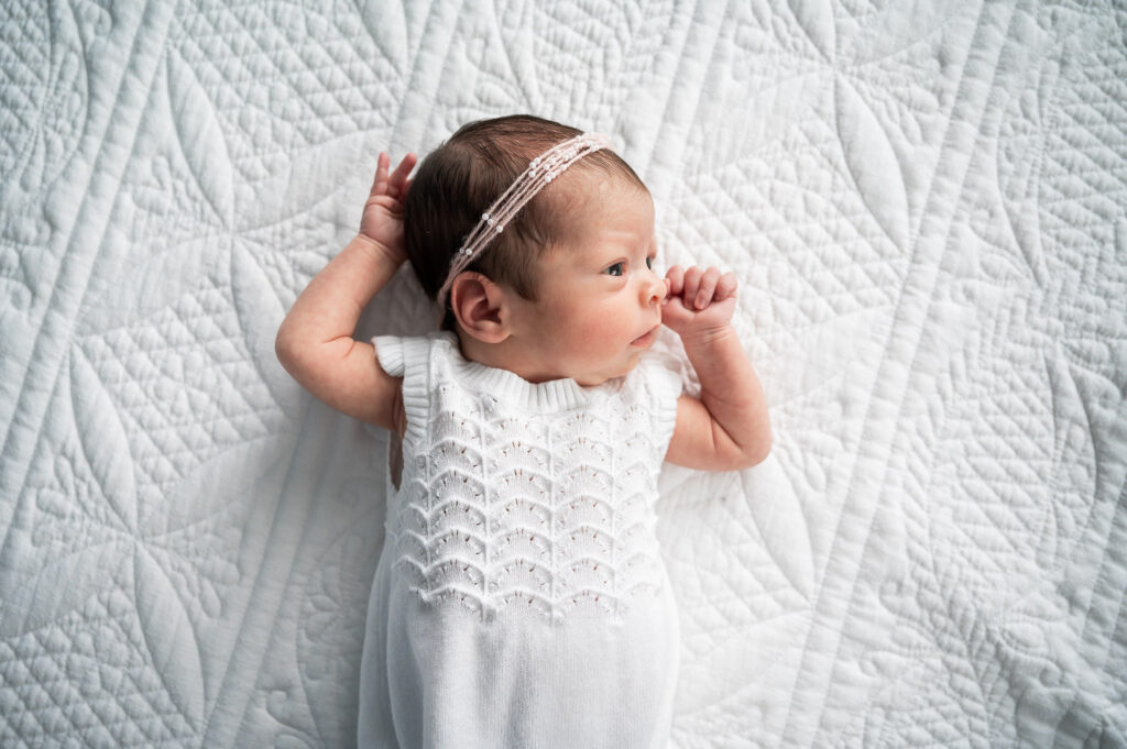 baby awake laying with her hands up next to her head wearing a pick headband and white knit outfit